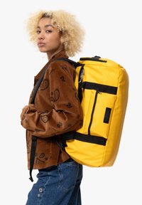 Mochila impermeable amarilla con detalles en negro, forma rectangular, bolsillo lateral y correas ajustables, llevada por una persona con una chaqueta marrón de estampado.