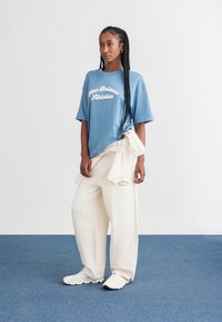 T-shirt oversized azul com "New Balance Athletics" em branco, combinado com calças de fato largas em tom crema. Ténis atléticos brancos.