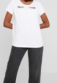 Camiseta blanca de manga corta con un gráfico de pestañas negras en el frente, cuello redondo y suave tela de algodón. Llevada con pantalones gris oscuro.