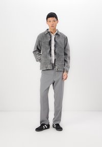 Gray denim jacket with buttons, worn over a plaid shirt. Light gray trousers and black sneakers with white accents complete the look.