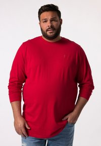 Rotes Langarmshirt aus Baumwolle mit Rundhalsausschnitt. Dezentes Logo auf der Brust. Kombiniert mit hellblauen Denim-Jeans.