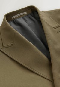 Olive zaļš bleizeris ar klasisku atloku un šuvju detaļām; iekšpuses odere ir pelēka ar smalku punktu rakstu.