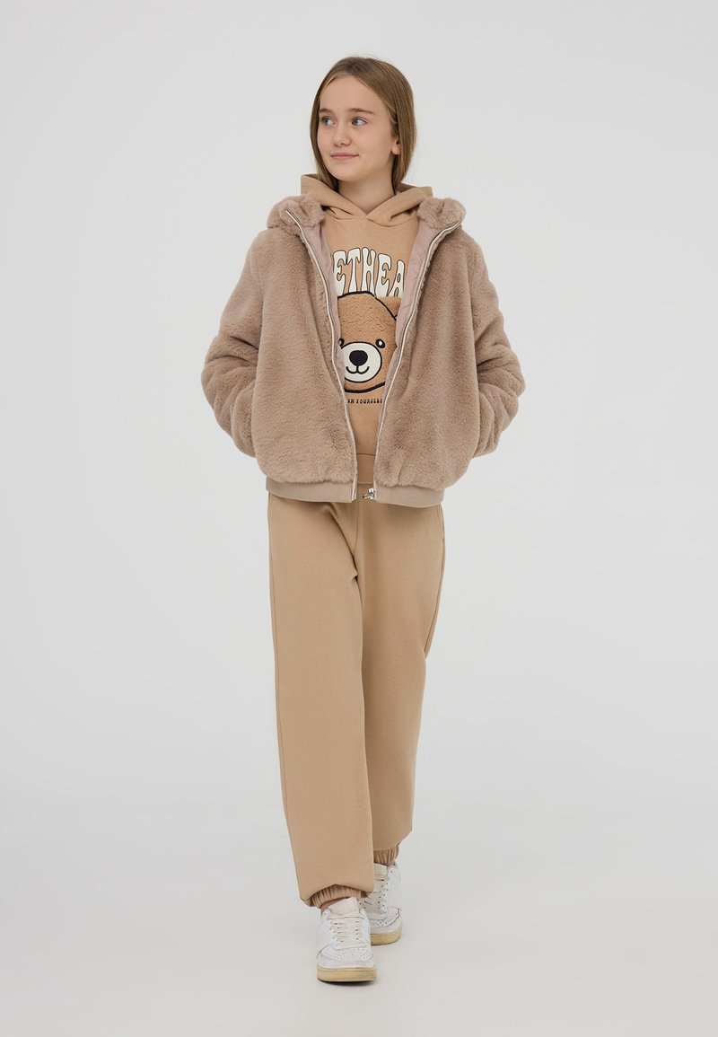 Giacca in finta pelliccia marrone con zip, felpa beige con grafica di orso, pantaloni jogger beige e sneakers bianche. Vestibilità rilassata, texture morbida, look casual.