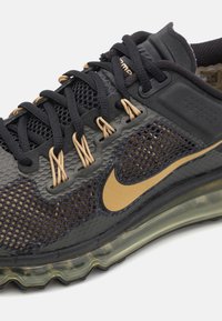 Juodos ir auksinės sportbačiai, pagaminti iš tinklinės medžiagos, su storomis juodomis raištelėmis, tekstūruotu tarpu ir ryškiu auksiniu Nike swoosh logotipu.