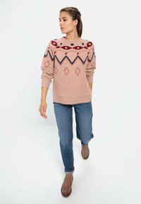 Strickpullover in Hellpink mit geometrischen Mustern in Rot, Schwarz und Creme. Kombiniert mit blauen Jeans und braunen Knöchelstiefeln.