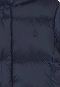 Marinblå pufferjacka med huva, med quiltade sektioner och en broderad logo i det övre vänstra hörnet. Slät, glänsande tygstruktur.