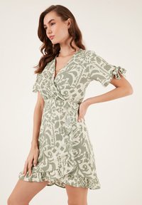 Wickelkleid in Hellgrün mit Tierdruck, kurzen Ärmeln, die an den Schultern gebunden sind, Rüschen am Saum und einem Knopfdetail an der Vorderseite.