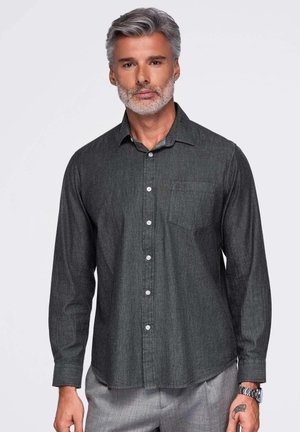 Homme aux cheveux gris et à la barbe, portant une chemise boutonnée gris foncé et un pantalon gris clair, debout devant un fond blanc.