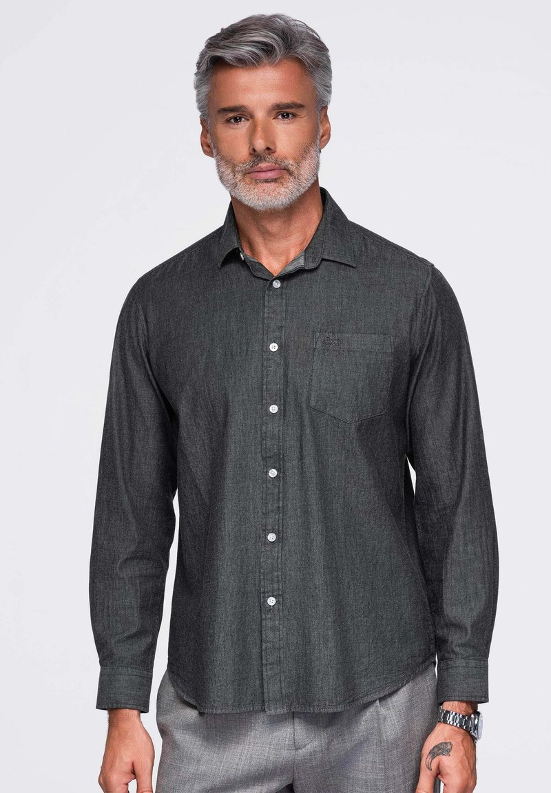 Homme aux cheveux gris et à la barbe, portant une chemise boutonnée gris foncé et un pantalon gris clair, debout devant un fond blanc.