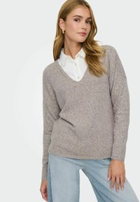 Pull taupe clair avec un col en V, à manches longues, au fit décontracté et en tricot texturé. Associé à une chemise blanche à col et un jean en denim clair.