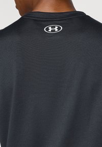 Chemise de sport noire en tissu texturé. Présente un col rond et un logo blanc au dos, près du col.