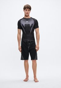 Μαύρο αθλητικό rash guard με κοντά μανίκια και υφασμένη υφή, σετ με μαύρο μαγιό σορτς που διαθέτει αντίθετες ραφές για διακριτικές λεπτομέρειες.