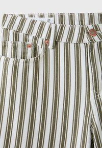 Gros plan d'un pantalon rayé avec des lignes verticales olive et blanches, une poche avant, des passants de ceinture et des rivets en cuivre près de la taille.
