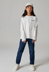 Valge pika varrukaga särk, millel on "ALLSAINTS" logo, sinised denim teksad, valged tennis jalanõud ja valge pesapallimüts logoga. Lihtne ja mugav disain.