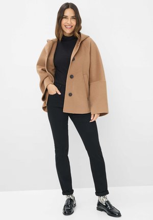 Bruine wolblendjas met een capuchon, voorzien van grote zwarte knopen, ruime mouwen en een ontspannen pasvorm, gedragen over een zwarte top en zwarte broek.