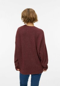 Višeslojni pulover tamnocrvene boje s okruglim izrezom i rebrastim rubom, izrađen od mekanog, teksturiranog materijala. Dugi rukavi s padanim ramenima.