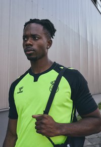 Maglia sportiva verde e nera con maniche corte, caratterizzata da un colletto polo, logo Hummel e emblema della squadra sul petto. Accenti laterali visibili.