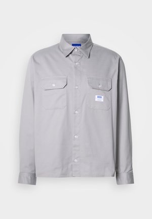 Camisa cinza de mangas longas feita de algodão, com colarinho abotoado, dois bolsos no peito e uma pequena etiqueta de marca no bolso esquerdo.