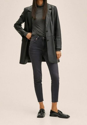 Persoon draagt een zwart leren blazer, donkergrijze coltrui, donkere skinny jeans met rafelige zomen en zwarte loafers, staand tegen een beige achtergrond.