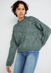 Maglione verde lavorato a maglia con un motivo a onde testurizzato e dettagli traforati, caratterizzato da un colletto rotondo e maniche leggermente oversize.