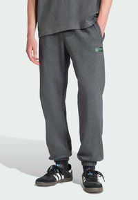 Grå mjukisbyxor i mjukt material med lös passform, elastiska muddar och en liten grön logotypapplikation. Bärs med svarta sneakers och strumpor med blå detaljer.