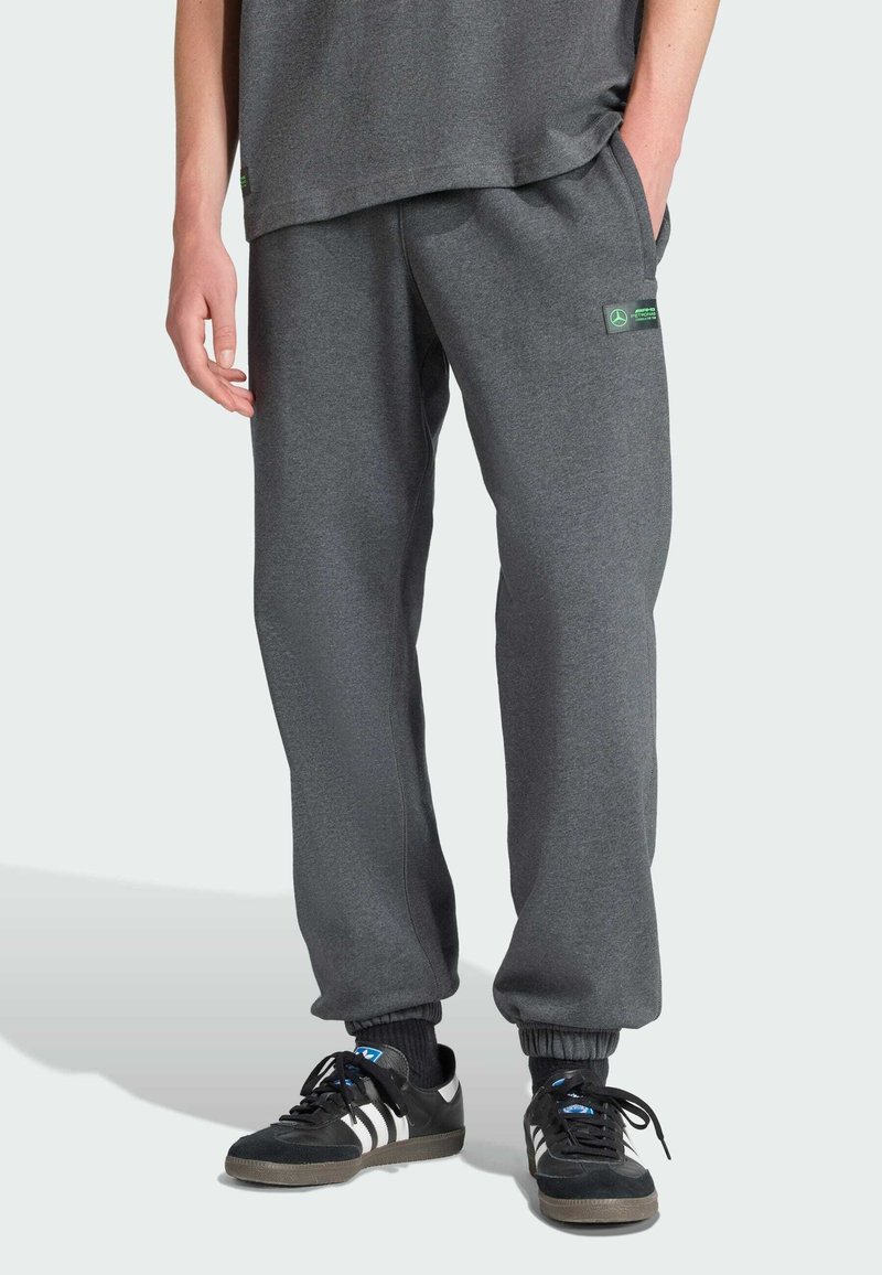 Grå mjukisbyxor i mjukt material med lös passform, elastiska muddar och en liten grön logotypapplikation. Bärs med svarta sneakers och strumpor med blå detaljer.