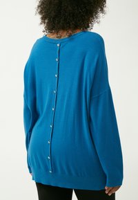 Übergroßer blauer Pullover mit einer weichen Textur. Verfügt über eine Knopfverschluss auf der Rückseite und ein geripptes Bündchen als zusätzliches Detail.