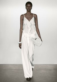 Weiße mehrschichtige Bluse mit Rüschen und Knöpfen, kombiniert mit lockeren weißen Hosen. Das Modell hält eine strukturiert gewebte Tasche und trägt flache Sandalen.