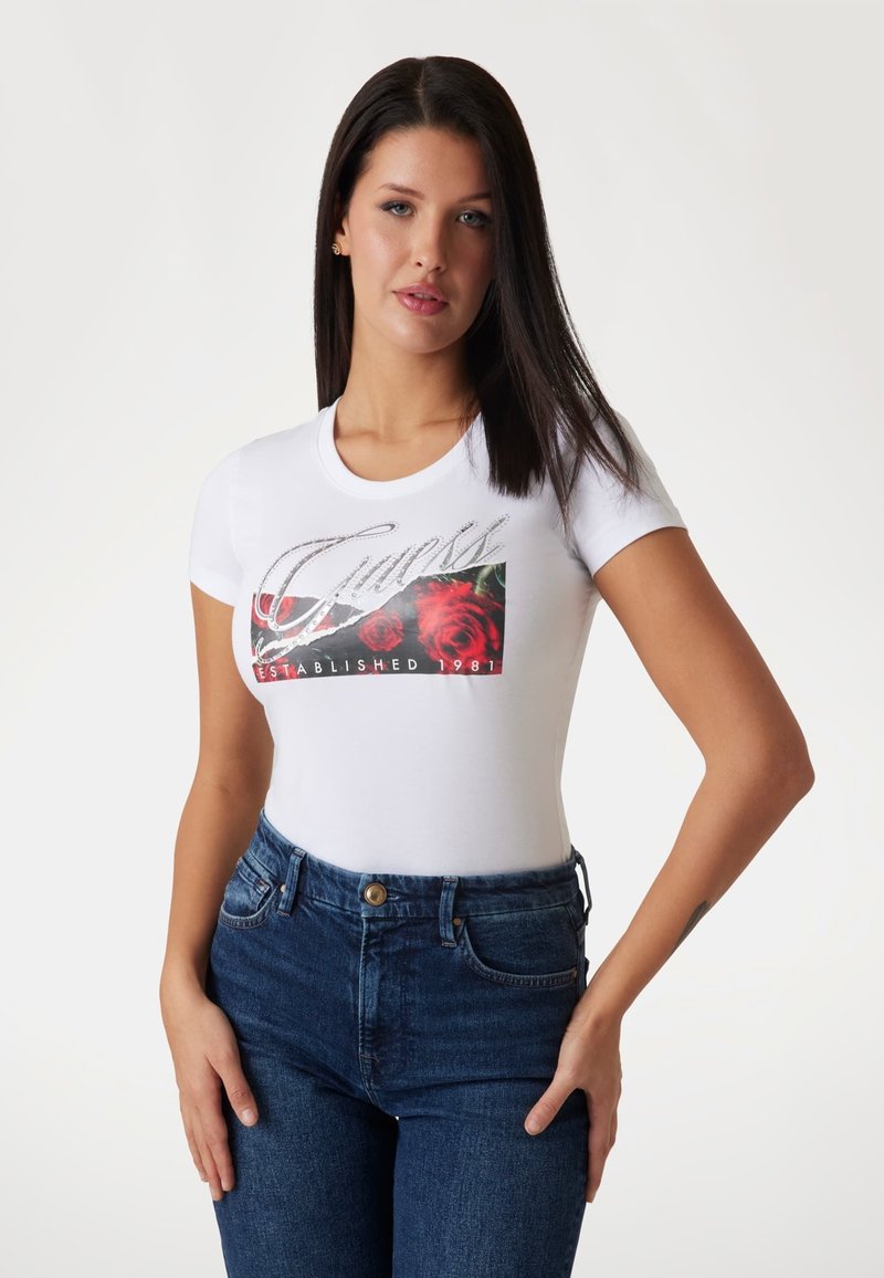 Biela tričko s krátkym rukávom a grafickým dizajnom, ktorý zobrazuje červené ruže a nápis "Guess Established 1981" v striebornom písmene. Tkanina sa zdá byť hladká.
