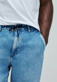 Jeans de mezclilla azul claro con un corte relajado, cintura con cordón y costuras en contraste. Acompañados de una camiseta blanca sencilla.