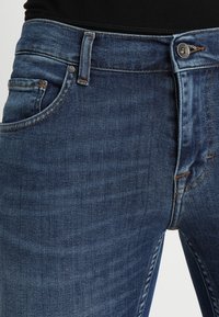Primer plano de una persona que lleva jeans de mezclilla azul de tiro medio con bolsillos frontales, botón y cinturas, combinados con una blusa negra.
