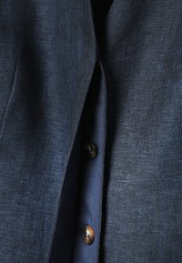 Primer plano de tela texturizada de chaqueta azul oscuro con dos botones marrones en la capa interior de la prenda.
