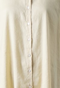 Camicia button-up beige chiaro realizzata in tessuto morbido, con chiusura frontale a bottoni bianchi e una vestibilità rilassata.