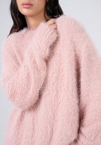 Lichtroze fluffy trui met lange mouwen, geribde boorden en een ruime pasvorm. Het materiaal lijkt zacht en gestructureerd, wat de warmte verhoogt.