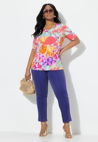 Multikleuren graphic top met korte mouwen, met flamingo- en geometrische patronen, gecombineerd met paarse tapered pants en gouden hakken.