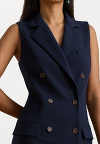 Marineblauwe mouwloze blazer met een dubbelgebreid ontwerp, voorzien van bruine knopen en een voorke pocket. Getailleerde pasvorm met een soepele textuur.