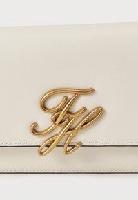 Αξεσουάρ από κρεμ δέρμα με χρυσά, πλεγμένα αρχικά "TH" και εμφανείς ραφές κατά μήκος των άκρων.