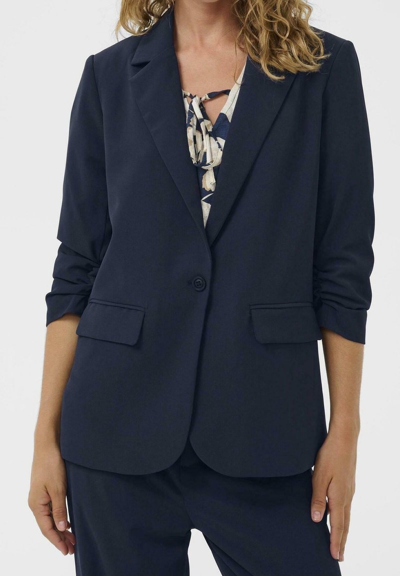 Femme portant un blazer bleu marine foncé avec les manches retroussées sur un chemisier à motifs avec un détail noué.