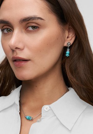 Frau mit türkisfarbenem Perlenhalsband und passenden Ohrhängern, gekleidet in weißes Hemd mit Kragen, mit langen braunen Haaren.