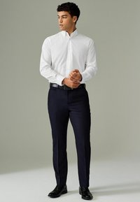 Chemise blanche à boutons, pantalon ajusté bleu marine foncé, ceinture noire et chaussures noires ; tissu lisse, coupe droite, détails minimalistes.