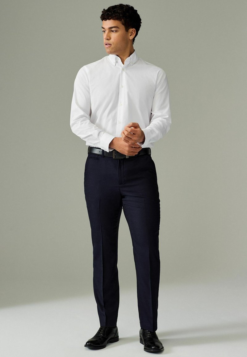 Chemise blanche à boutons, pantalon ajusté bleu marine foncé, ceinture noire et chaussures noires ; tissu lisse, coupe droite, détails minimalistes.