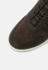 Gros plan sur l’avant d’une basket vert foncé et marron avec des panneaux en cuir et en daim, des lacets ronds marron et une semelle blanche.