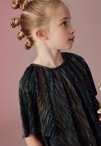 Schwarzes Kleid mit metallischen, schillernden Streifen. Verfügt über weiche, ausladende Ärmel und einen runden Ausschnitt. Der Hintergrund ist ein zartes Rosa.
