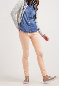 Jean skinny pêche clair associés à une chemise en denim et un cardigan gris, avec un motif texturé sur les manches et des chaussures à enfiler métalliques.