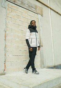 Veste rembourrée gris clair avec une partie supérieure noire, associée à un pantalon cargo noir et des baskets noir et blanc. Fond de mur texturé.