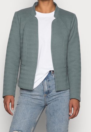Persoon die een grijze gestructureerde jas met rits draagt over een wit T-shirt en lichtblauwe spijkerbroek met een gescheurde knie, staand tegen een effen achtergrond.