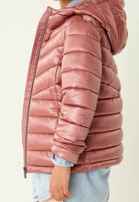 Veste rembourrée rose avec capuche, dotée de sections matelassées horizontales, d'une fermeture à glissière complète et de poignets élastiques. Portée par-dessus une chemise en denim clair.