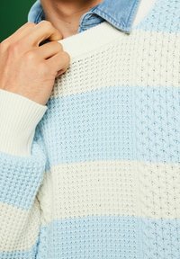 Hellblau-weiß gestreifter Strickpullover mit strukturiertem Muster, mit gerippten Bündchen und Ausschnitt. Über einem Jeanshemd getragen.