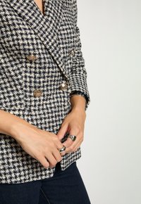 Primo piano di mani che allacciano un blazer bianco e nero a motivo houndstooth con bottoni dorati, indossando anelli d'argento e jeans in denim scuro.