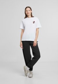 Hvit bomull t-skjorte med korte ermer som har et lite brodert design, kombinert med svarte joggebukser og lyse treningssko.
